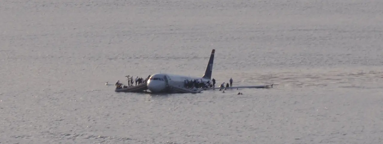 US Airways Flight 1549 floating low in the Hudson River, passengers standing on both wings and in inflatable rafts alongside the fuselage.