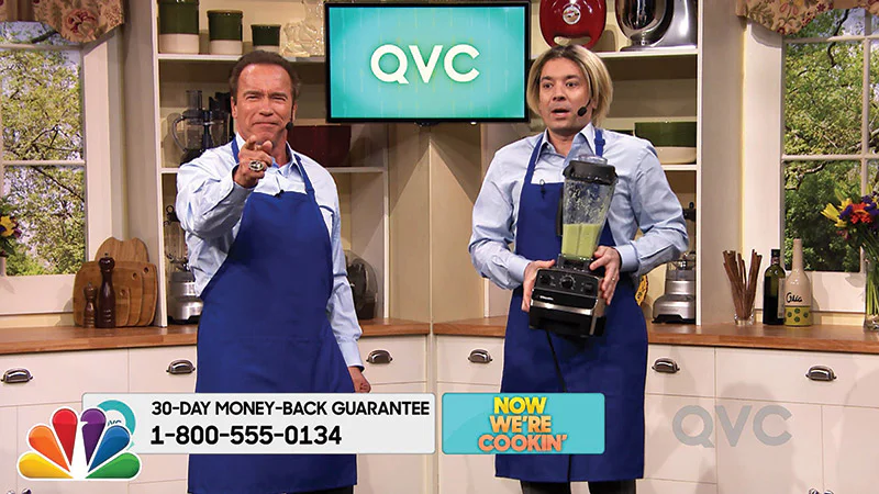 Two hosts in blue aprons on a QVC-style set, one holding a blender, in a late-night comedy spoof of teleshopping