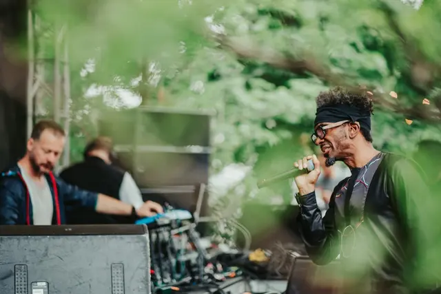 Musician singing into a microphone beside a producer at a mixing desk, outdoors among green trees