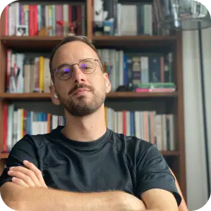 Portrait of Niko, a bearded man in glasses and a dark t-shirt, arms crossed in front of a bookshelf