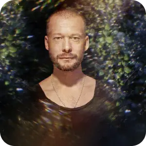 Portrait of Sebastian, a bearded man in a dark top, framed by dark foliage