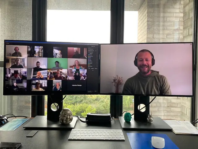 A host on a video call, smiling in headphones on one monitor, with a grid of participants on the other