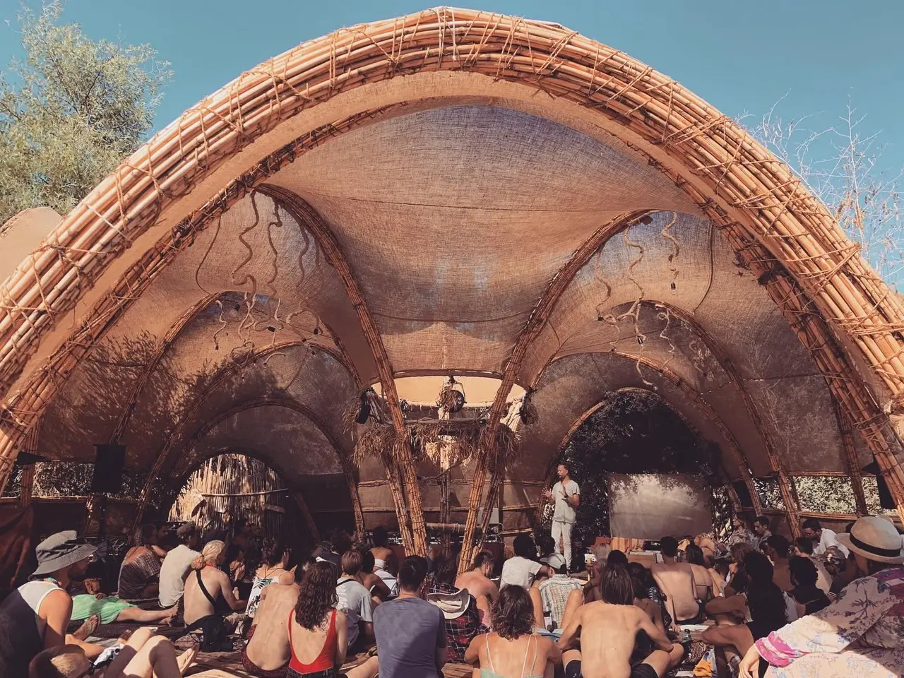 Speaker addressing a seated crowd under a large woven bamboo and fabric dome at an outdoor festival
