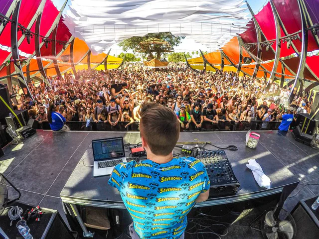 View over a DJ's shoulder at a laptop and mixer, facing a festival crowd with raised hands under a colorful canopy