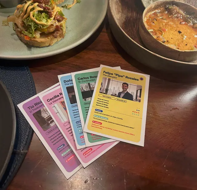 Fanned-out character cards for a family business game, printed in Spanish, on a restaurant table beside plates of food