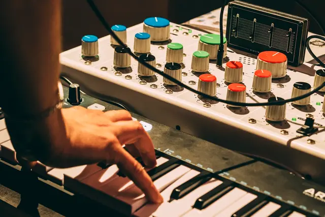 Hand playing a small keyboard next to a synthesizer with colorful knobs and patch points