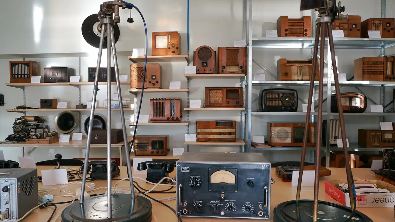 Museum room of vintage wooden radios on shelves, with two microphone stands and a measuring instrument on a table