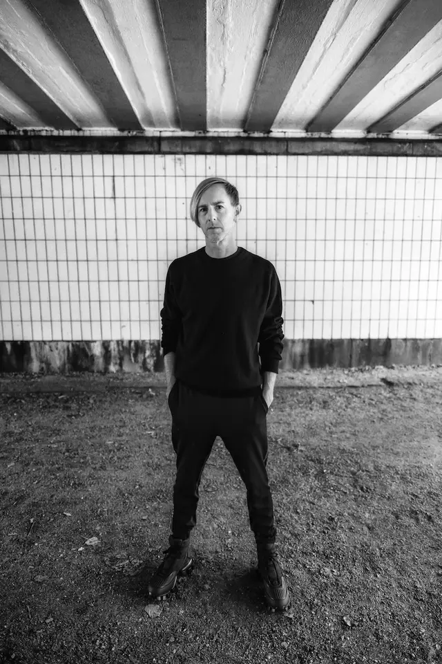 Black-and-white portrait of Richie Hawtin in dark clothes, standing under a concrete bridge in front of white tiles