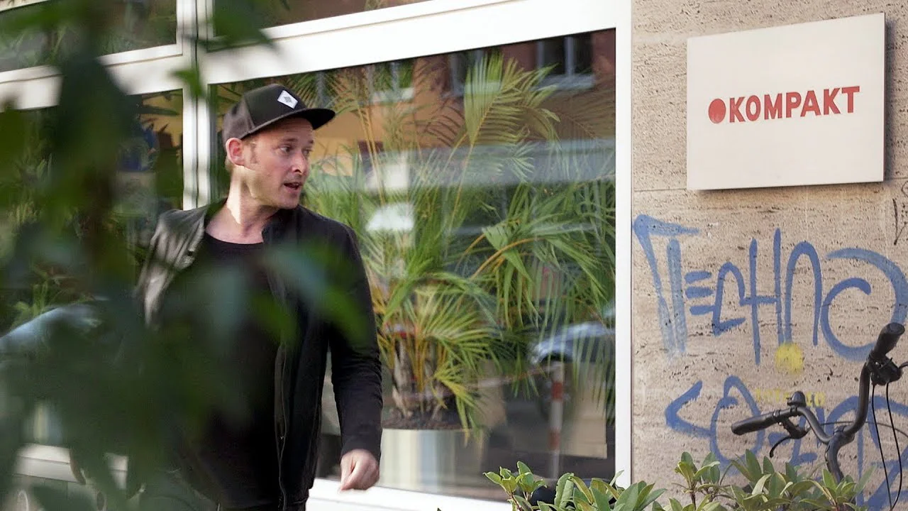 Michael Mayer in a cap and dark jacket outside a building with a Kompakt sign