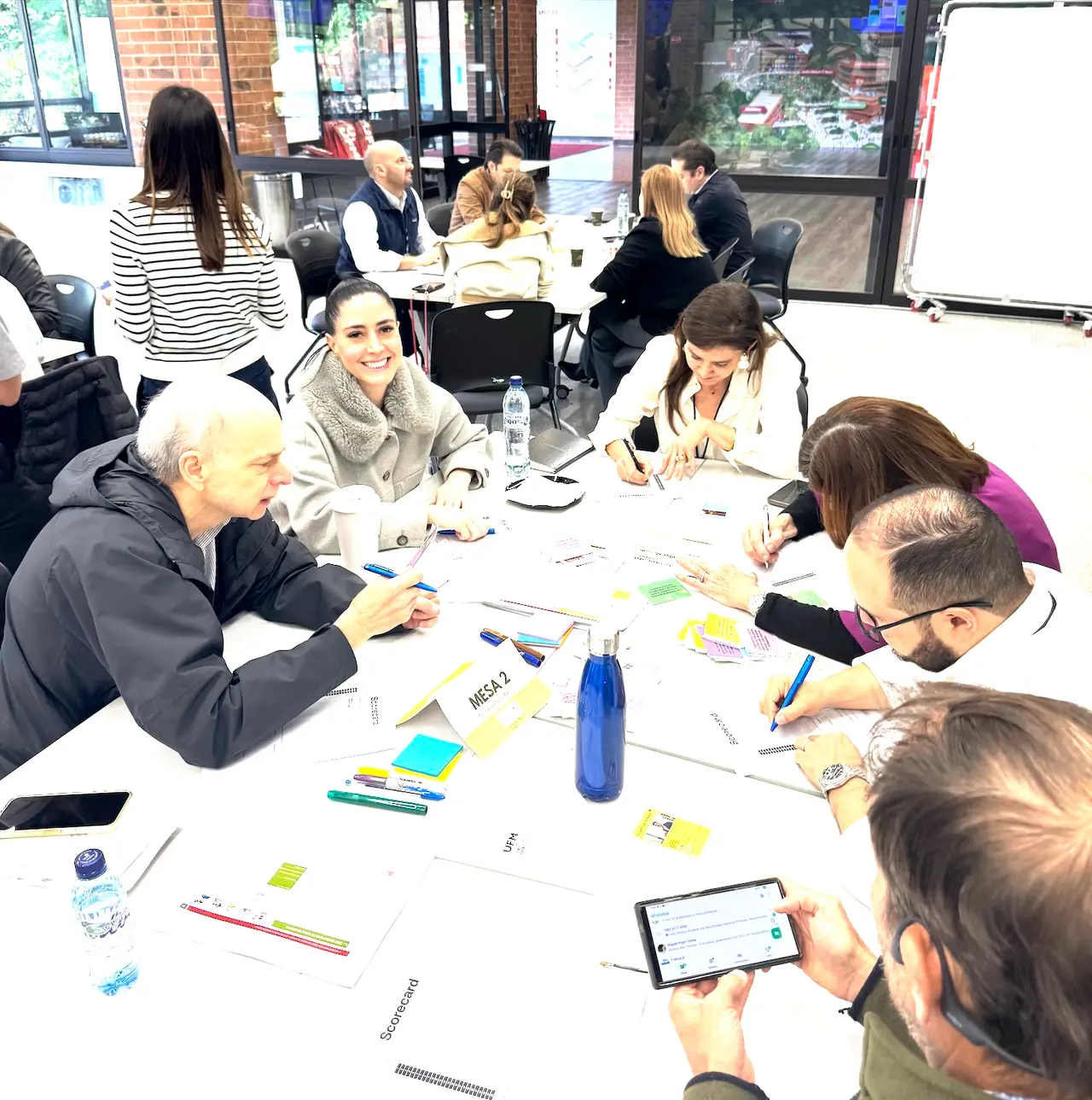 Workshop participants at a table, writing on scorecards among character cards and sticky notes, with a tablet in hand