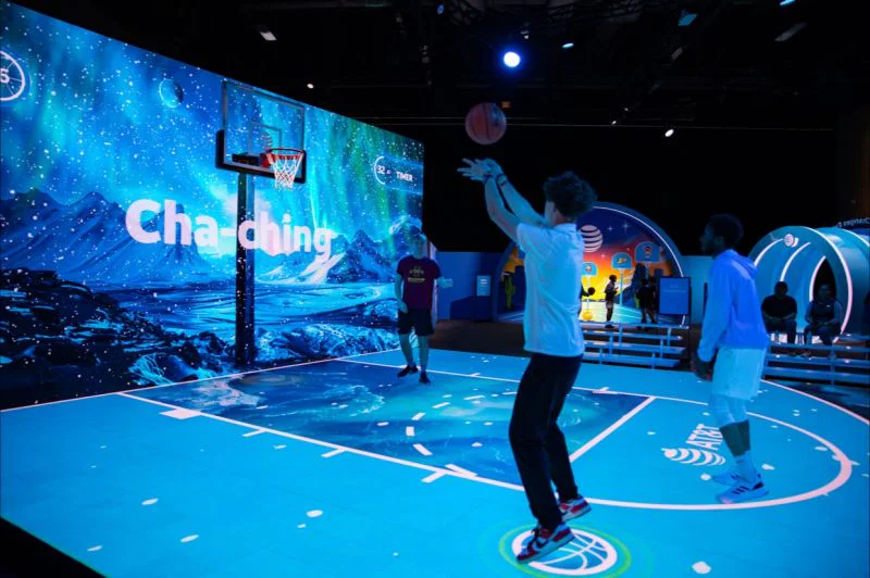 A player shoots at a hoop on a glowing projected basketball court, with Cha-ching on a huge screen of mountains and northern lights