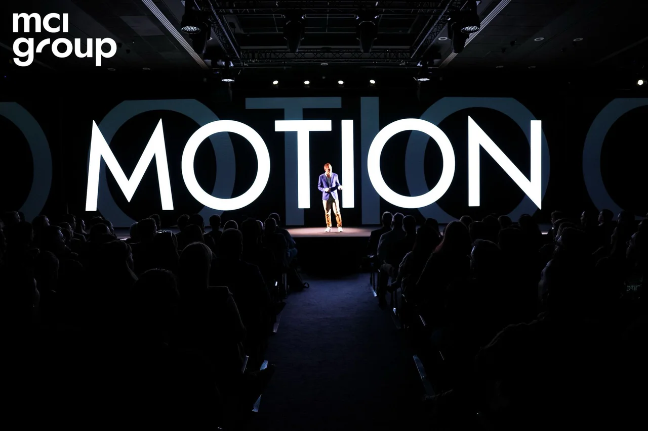 A speaker alone on a dark stage in front of the word MOTION in giant letters, the audience in silhouette