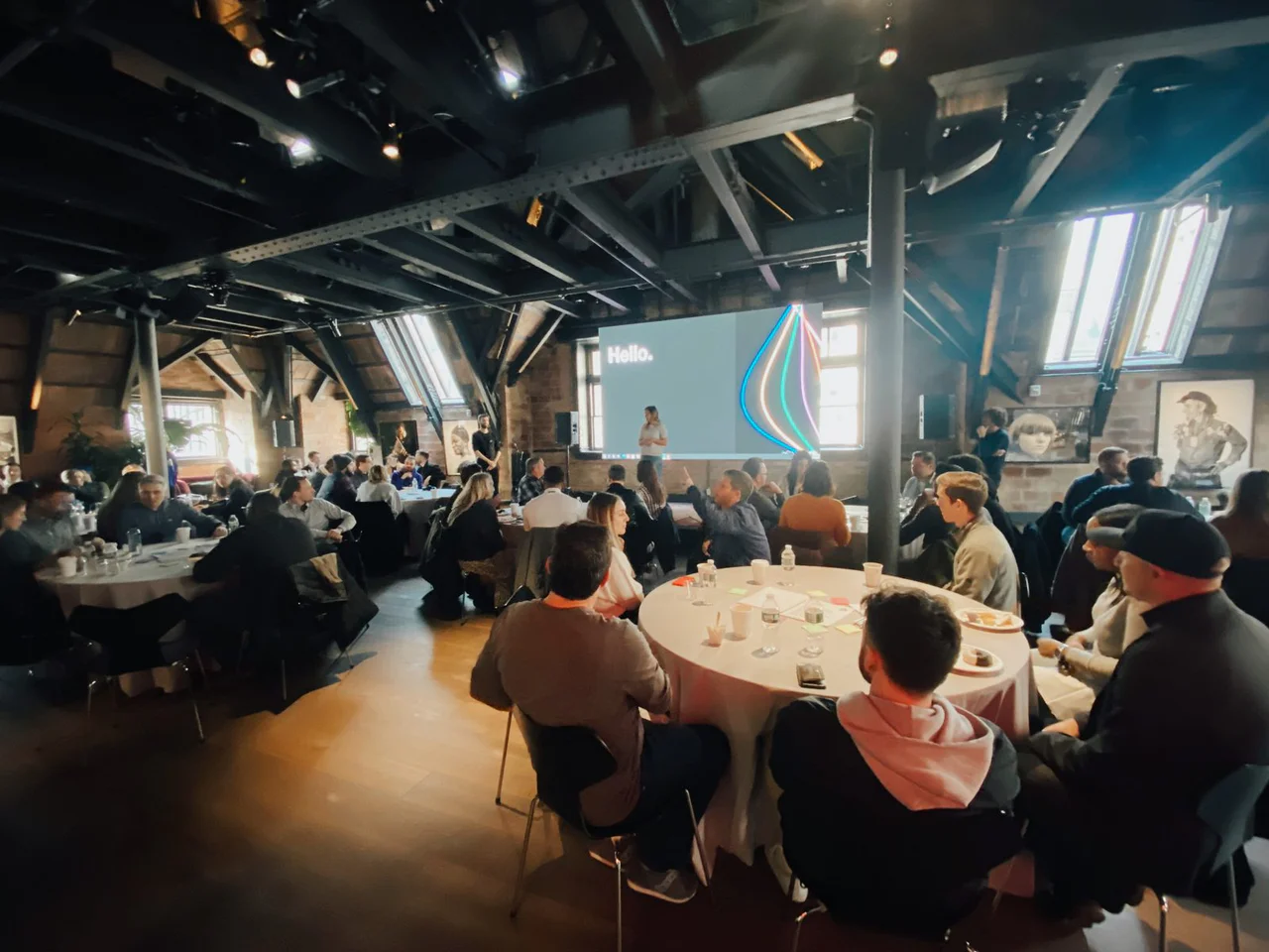 The hall in low light, a big screen reading Hello at the far end and tables of people seen from behind
