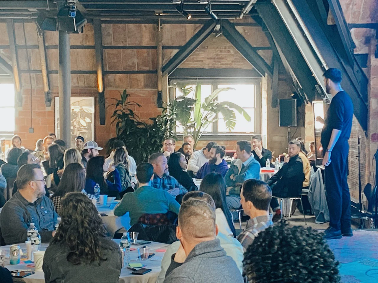 Round tables in a sunlit loft with exposed beams, the team seated and turned towards a host standing at the side