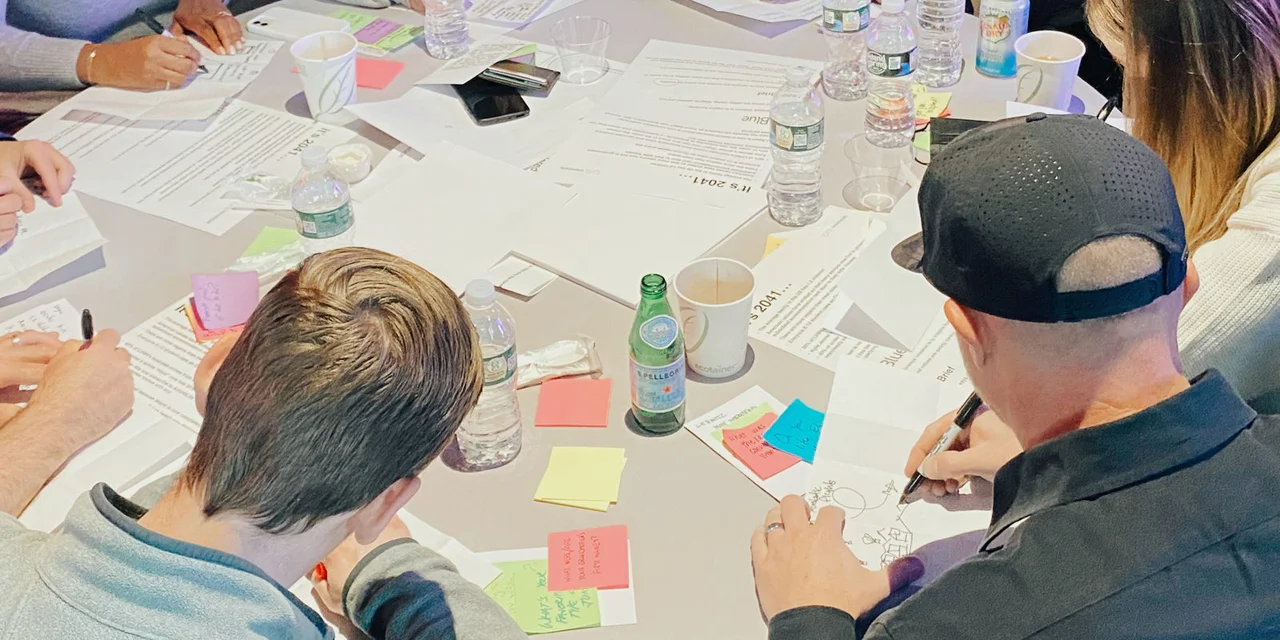 Hands writing on worksheets and sticky notes across a round table, water bottles and coffee cups between them