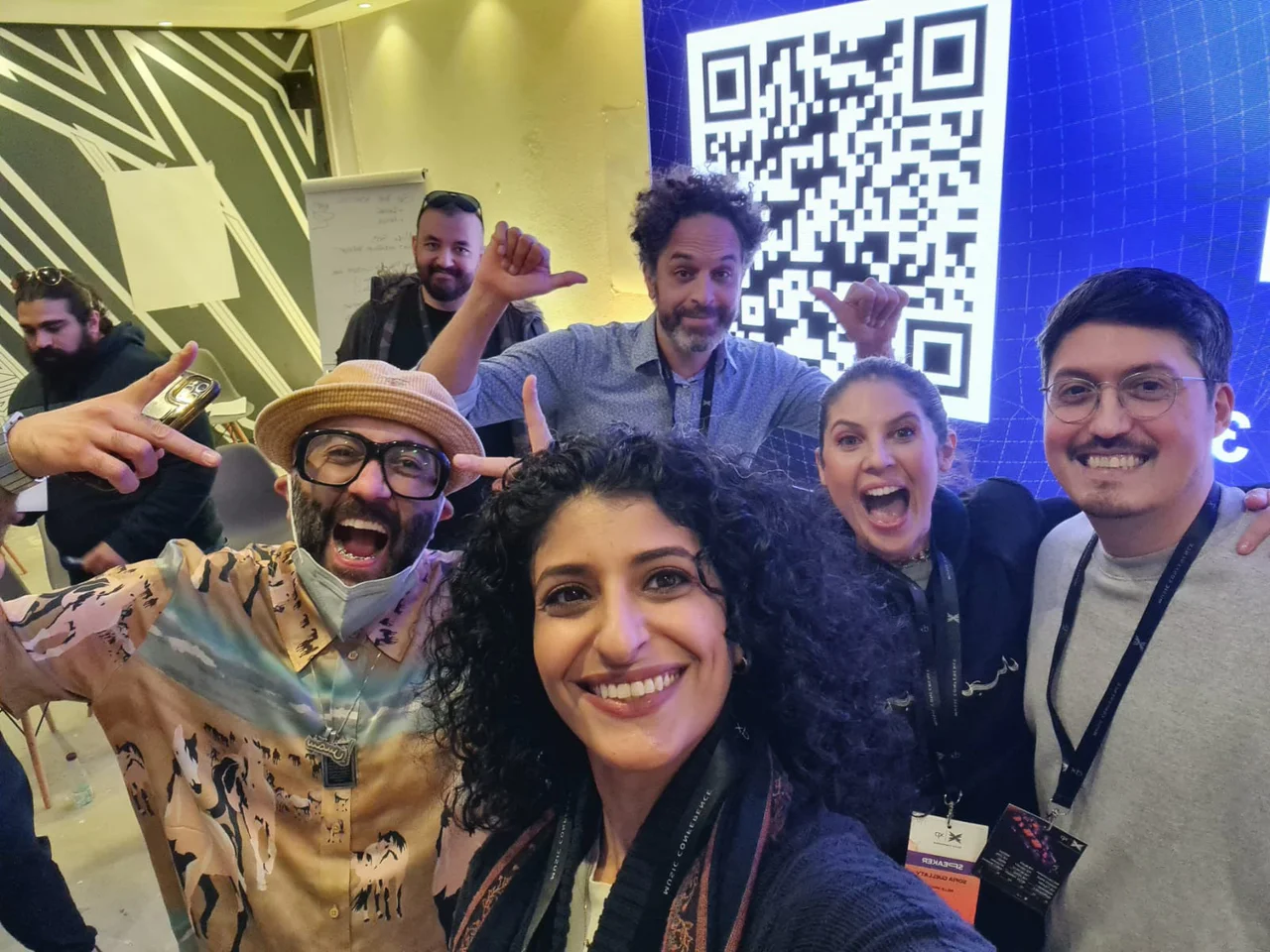 A selfie of the hosting crew in front of a QR code on the big screen, grinning, pointing and pulling faces