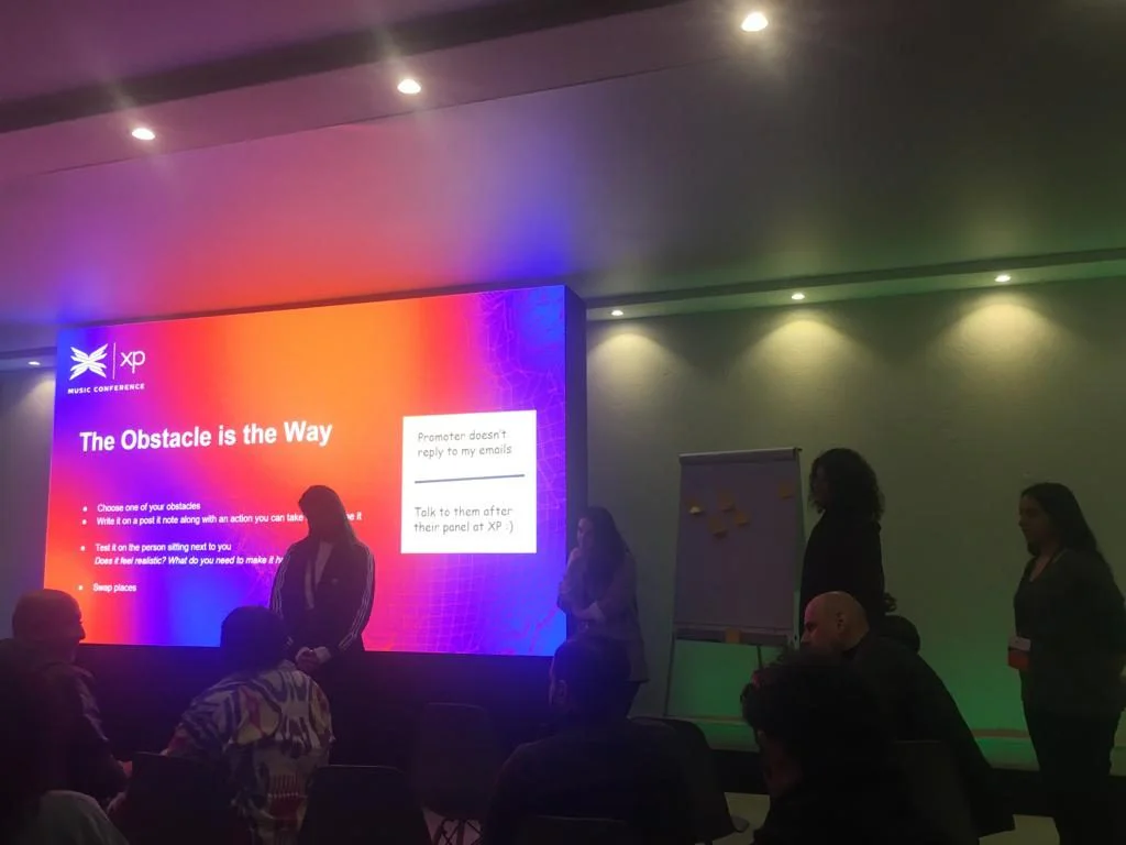 A workshop room in low light, a big screen reading The Obstacle is the Way, flip charts at the side and attendees seen from behind